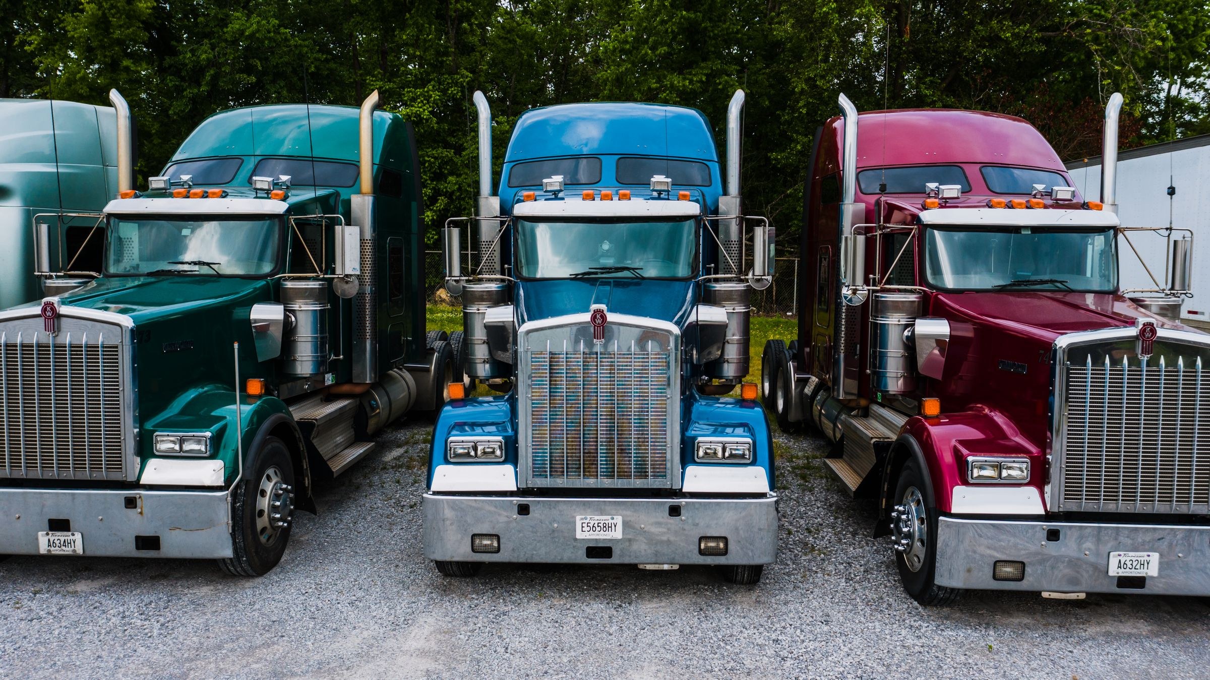 Green, blue and burgundy conventional semi trucks parked in a gravel yard with green trees