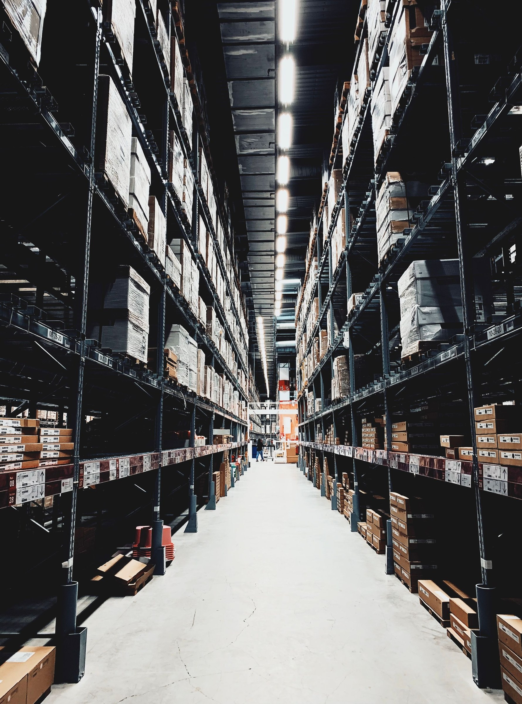 Tall pallet racks of wrapped cartons flank a long warehouse aisle beneath overhead lights