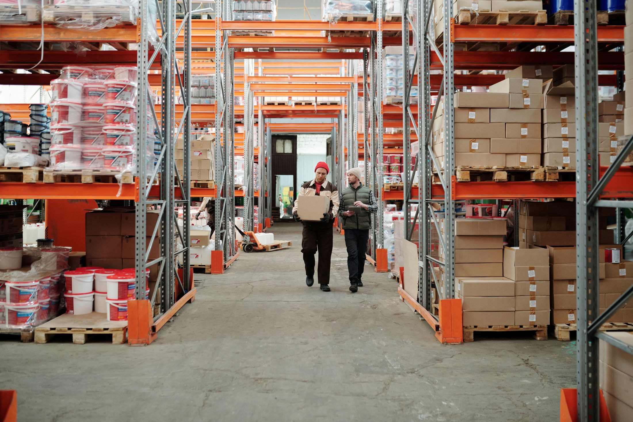 One worker carries a carton while another checks a tablet between orange warehouse pallet racks